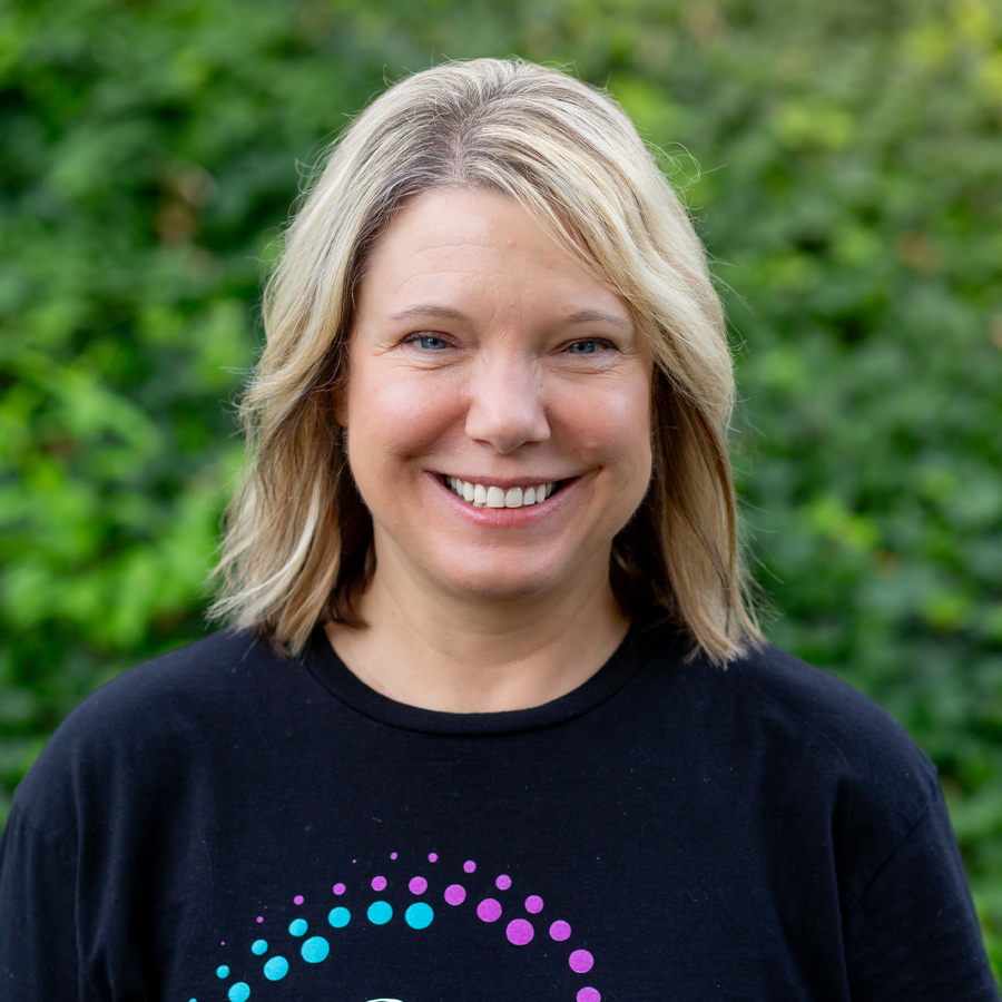 Blonde woman smiling, wearing a black shirt with a colorful design, standing outside near greenery.