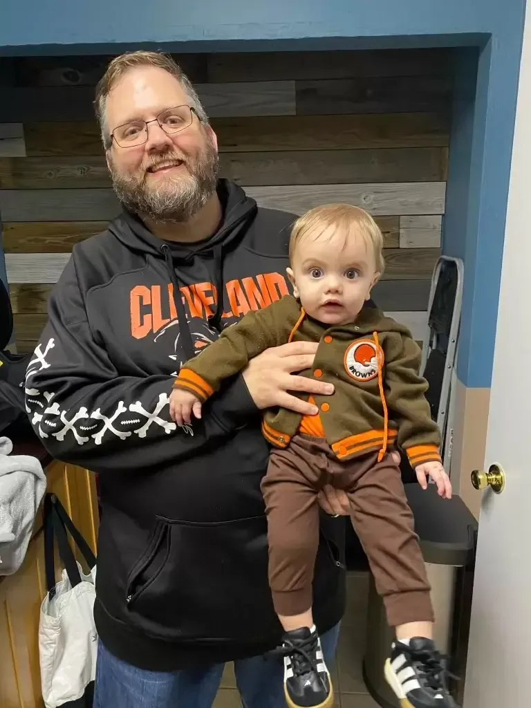 Man in a Browns hoodie holding a baby in a Browns jacket and pants. They are smiling.