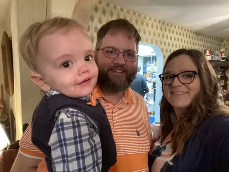 A family of three smiling at the camera: a baby, a man with a beard, and a woman with glasses.