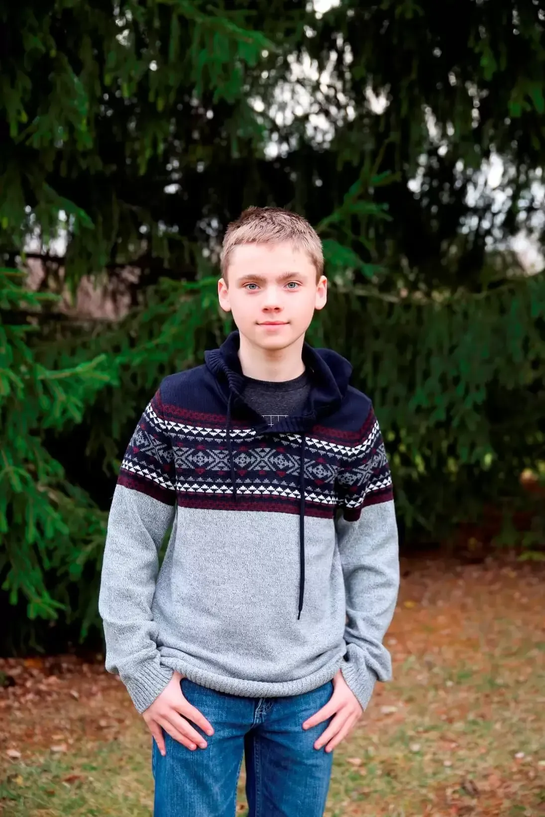 Teen boy with hands in pockets stands in front of a green tree. He wears a patterned sweater and jeans.