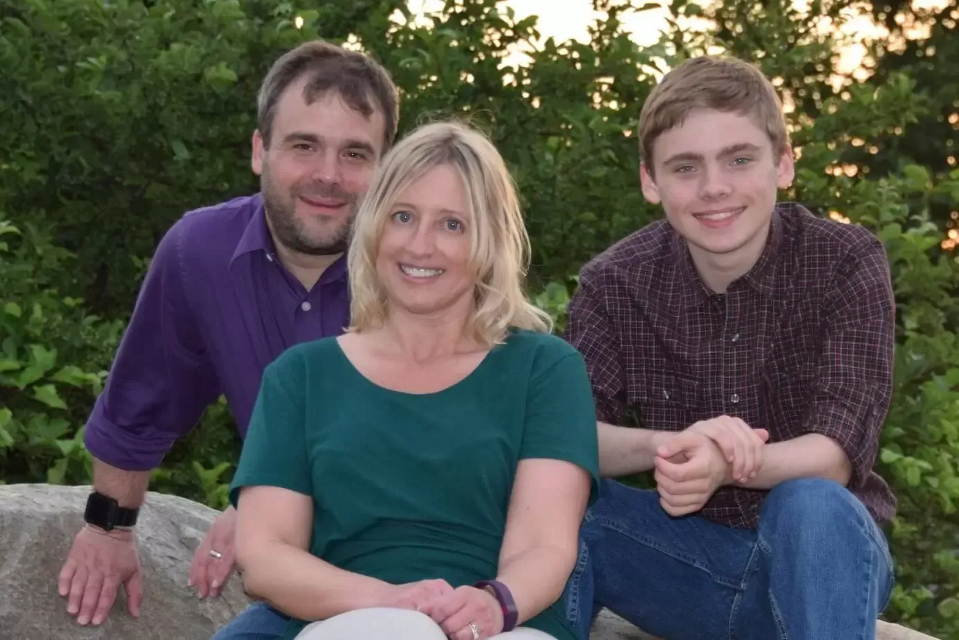 Family of three smiling outdoors; man in purple, woman in green, and teen in a button-down shirt.