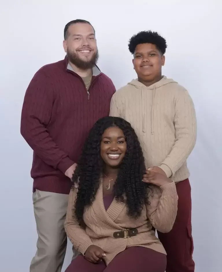 Family of three smiles in a studio: man in burgundy sweater, woman in tan sweater, and teen in a tan hoodie.