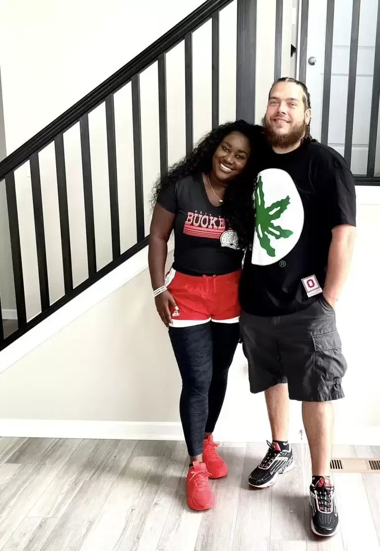 Smiling couple poses together. Woman in red and black outfit, man in black shirt with green design, and gray shorts.