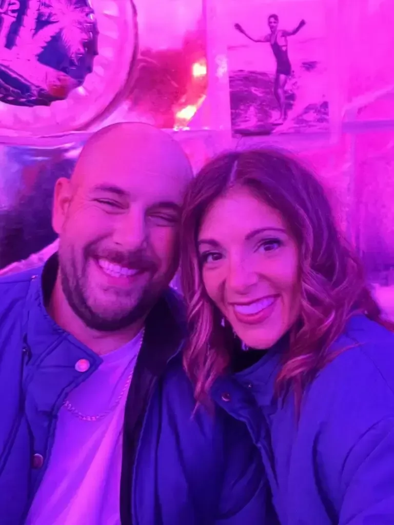Couple smiling for a selfie, illuminated by pink light, wearing blue jackets.