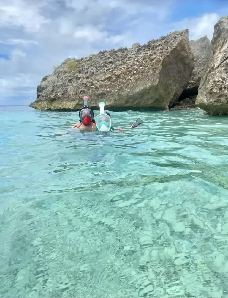 Two people snorkeling in clear turquoise water near rocky cliffs under a cloudy sky.