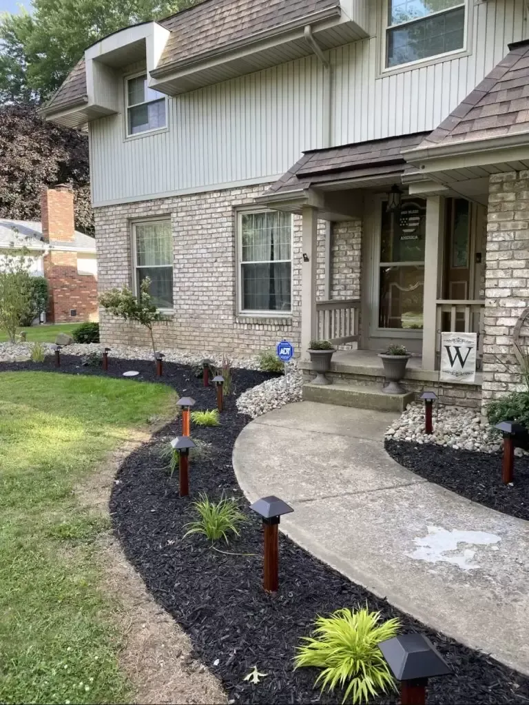 House exterior with a stone facade, concrete walkway, and landscaped flower beds with lighting.