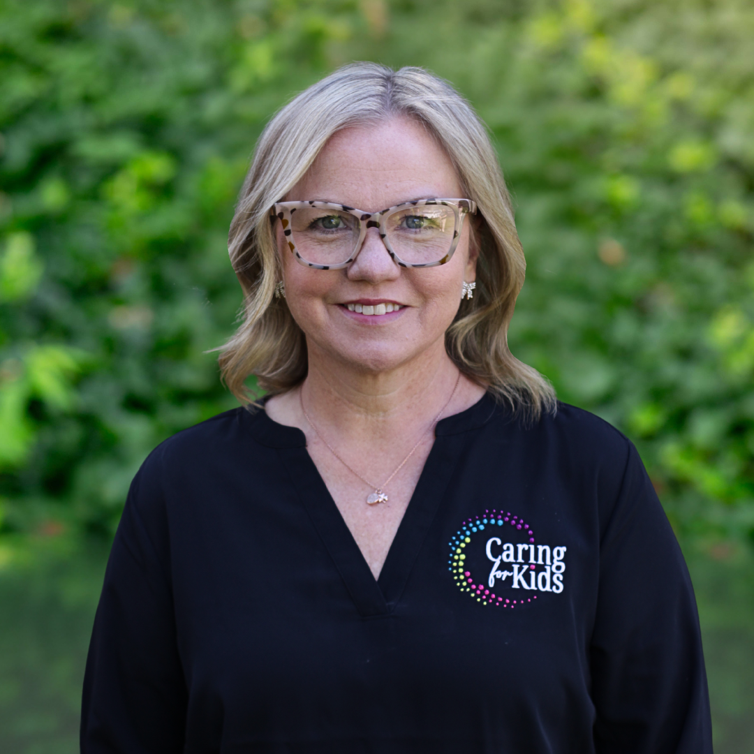 Woman with blonde hair, glasses, and earrings smiles, wearing a black speckled sweater, against a white background.