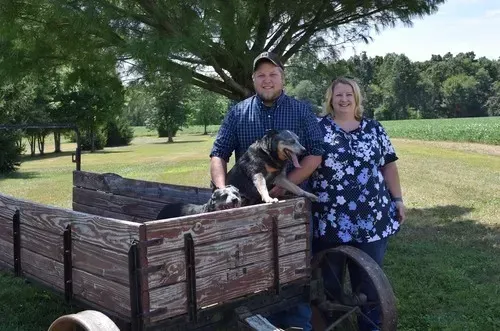 Couple and two dogs pose in a wooden wagon outside; man holds a dog, woman smiles.