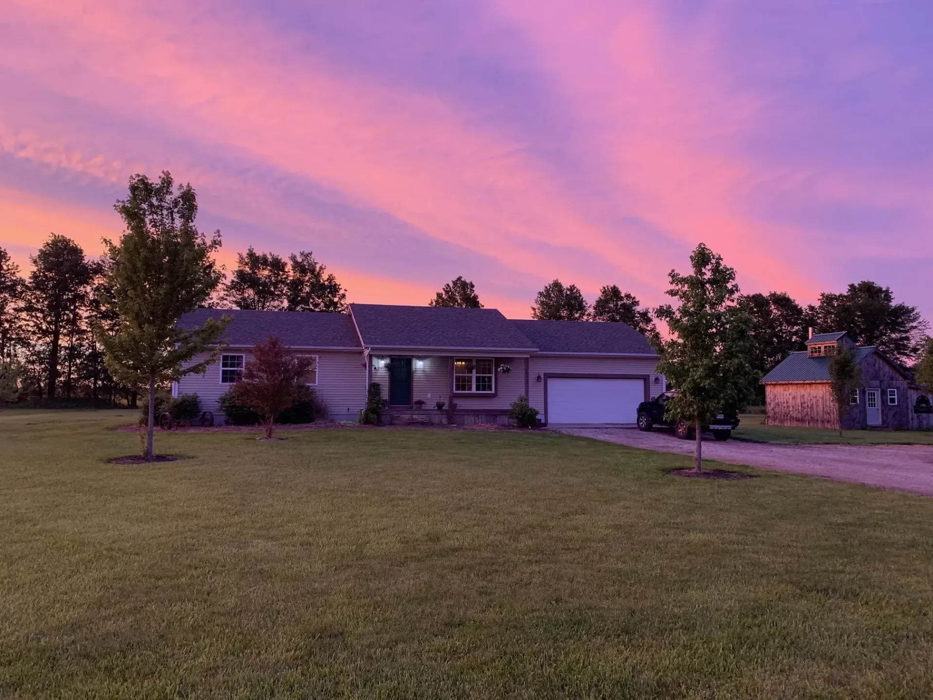 House at dusk with a pink and purple sunset, a green lawn, and a small outbuilding.