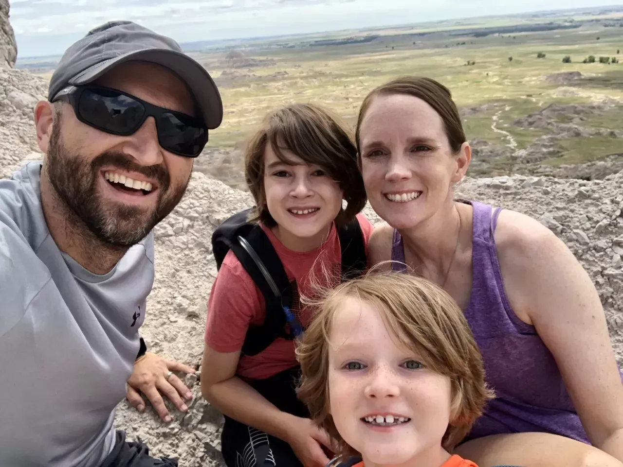 Family selfie smiling outdoors, man in sunglasses, woman, two boys with a scenic background.