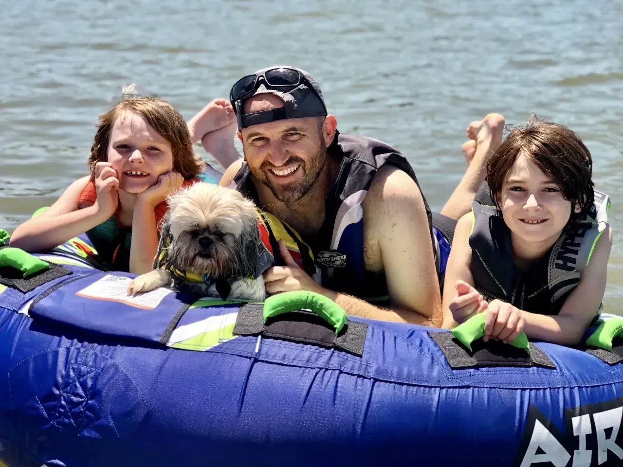 Family and dog on a blue inflatable tube on water, smiling. Sunny day.
