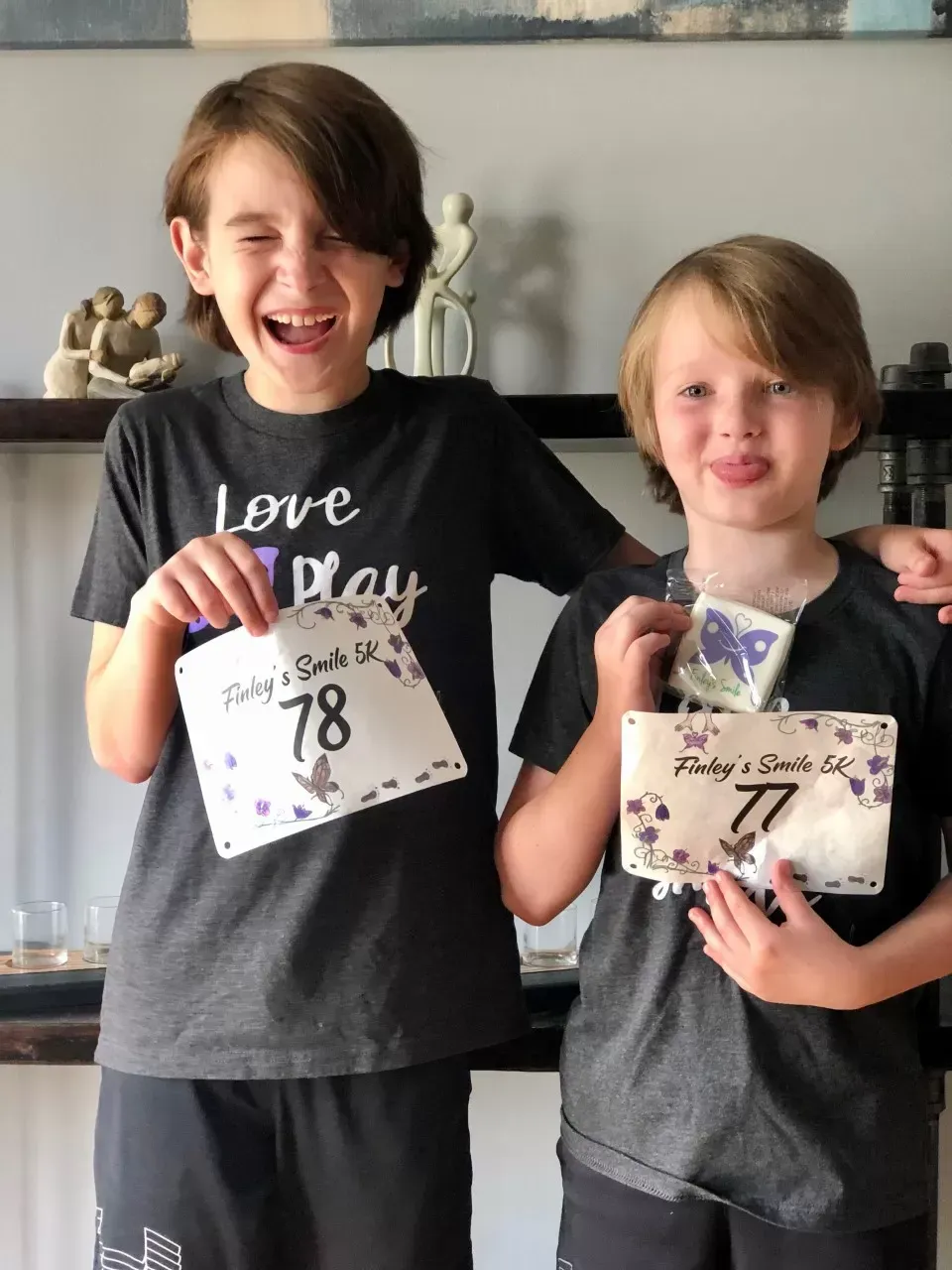Two boys, one laughing, the other sticking out their tongue, holding handmade race bibs in a home setting.
