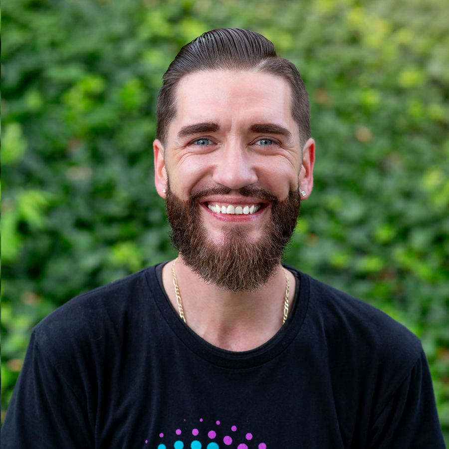 Smiling man with beard and slicked-back hair in front of green foliage; wearing black shirt with graphic.