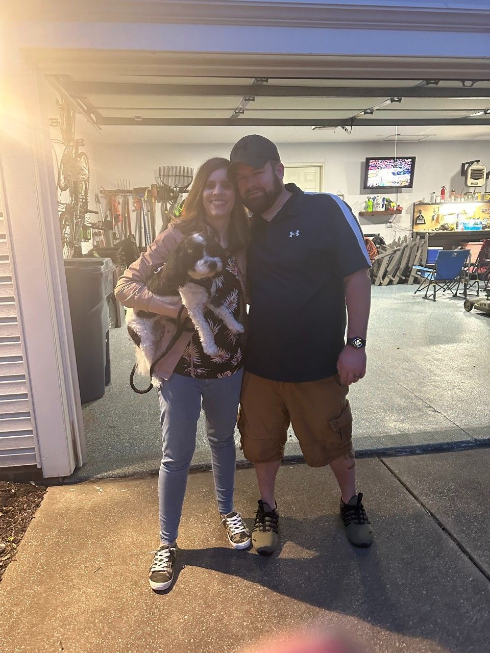Woman holding dog with man, standing in front of open garage.