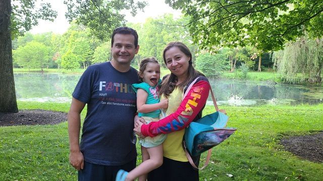 Family of three smiles by a pond. Father wears a blue shirt, mother a yellow and red jacket. Child is in center.