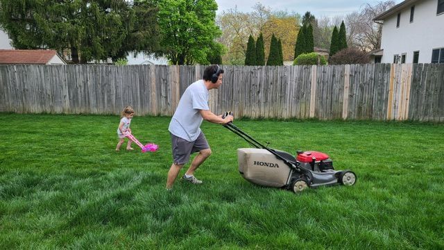 Man mowing lawn with a child following behind in a backyard, sunny day.