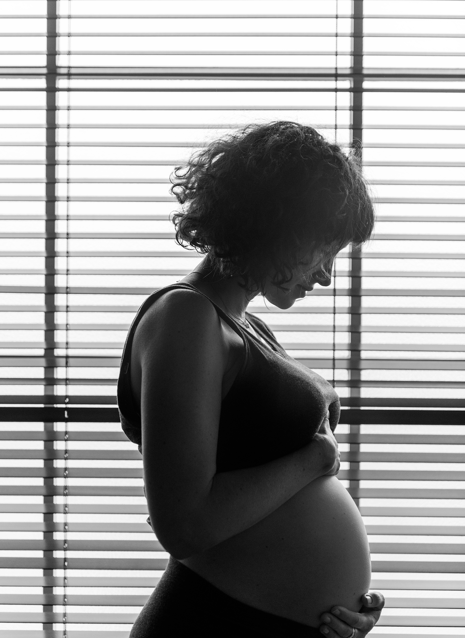 Pregnant woman in silhouette, holding her belly, in front of a window with blinds.
