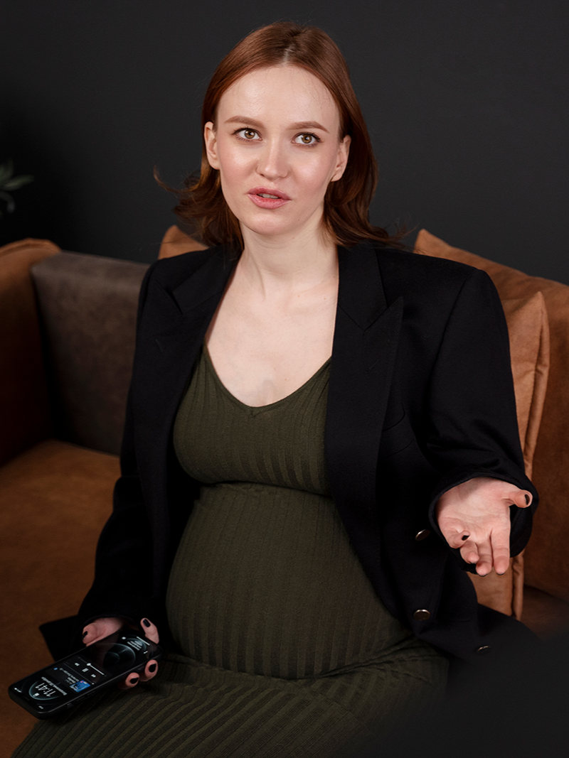 Pregnant woman in green dress and black blazer on a couch, holding ultrasound scan, with questioning expression.