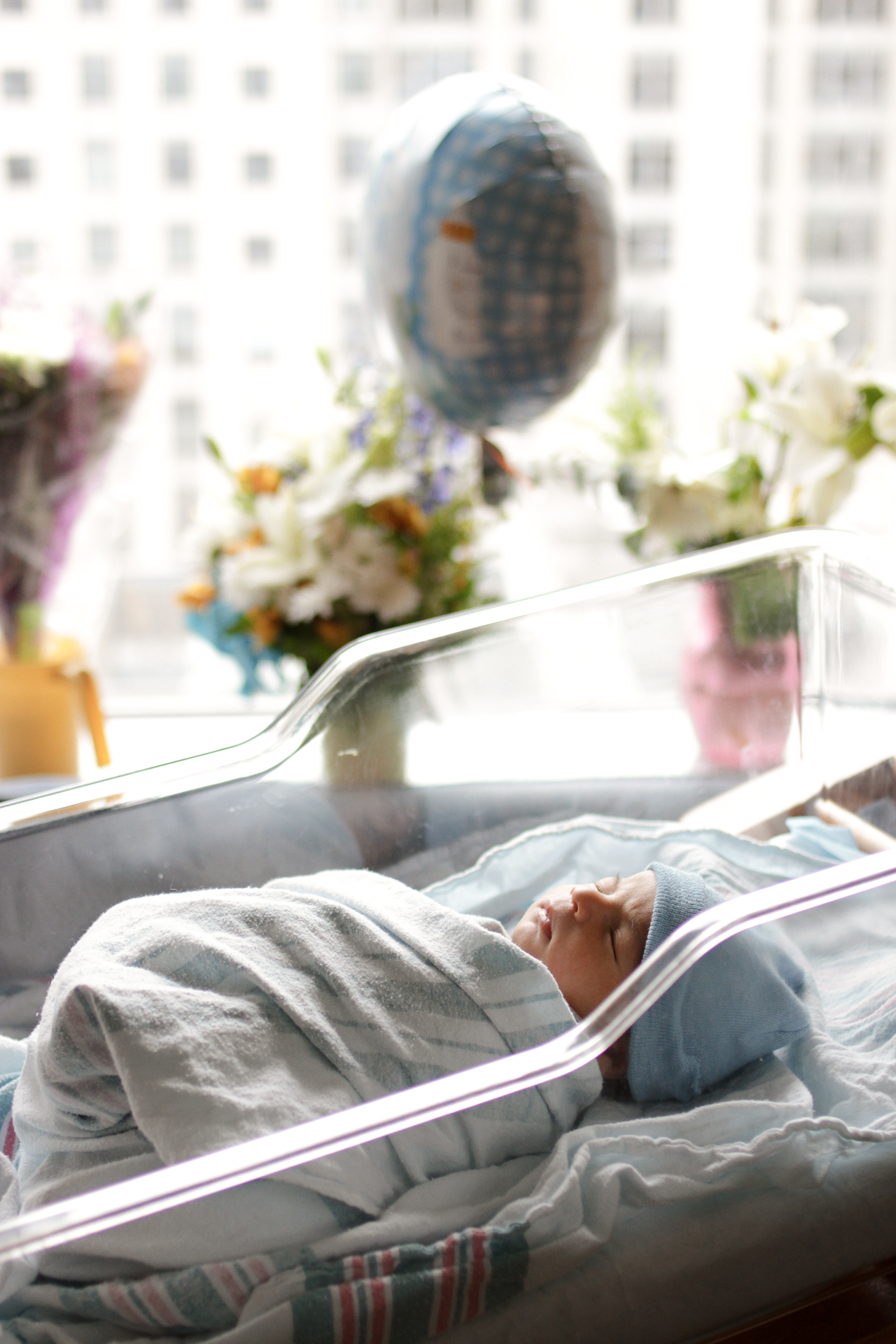 Newborn baby swaddled in a hospital bassinet, flowers and balloon visible in the background.