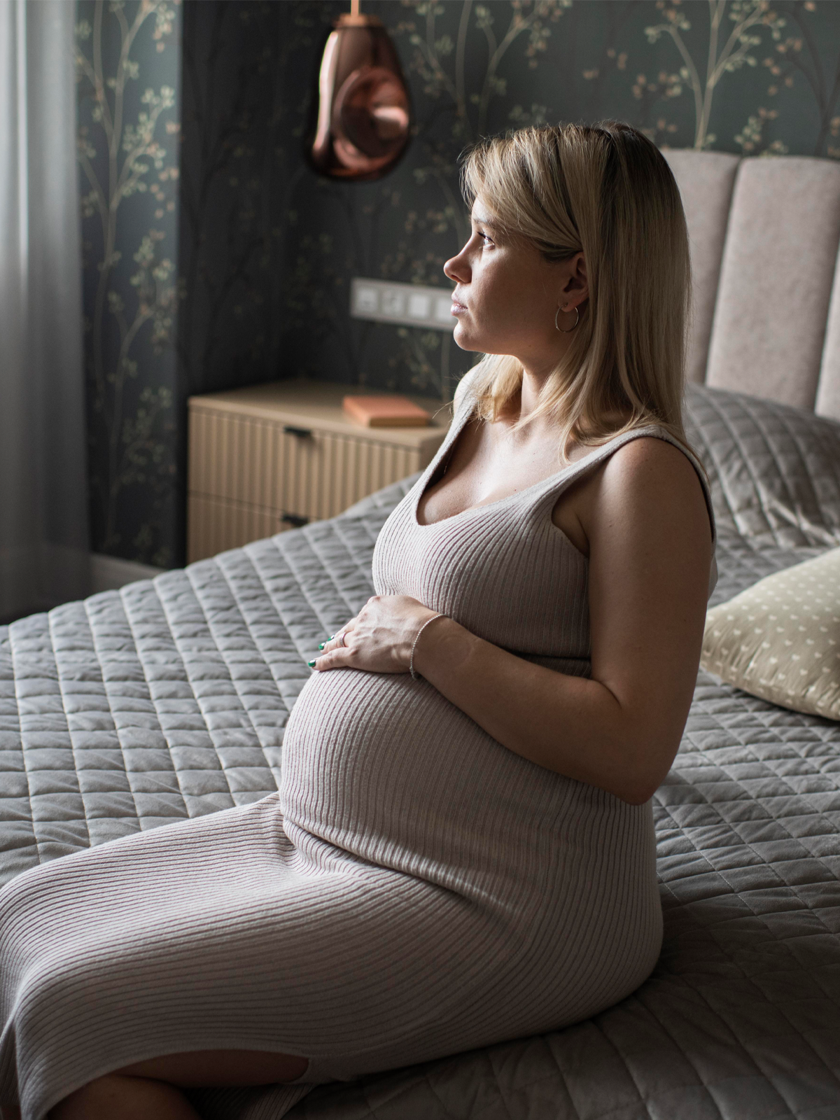 Pregnant woman in a beige dress, seated on a bed, looks out a window.