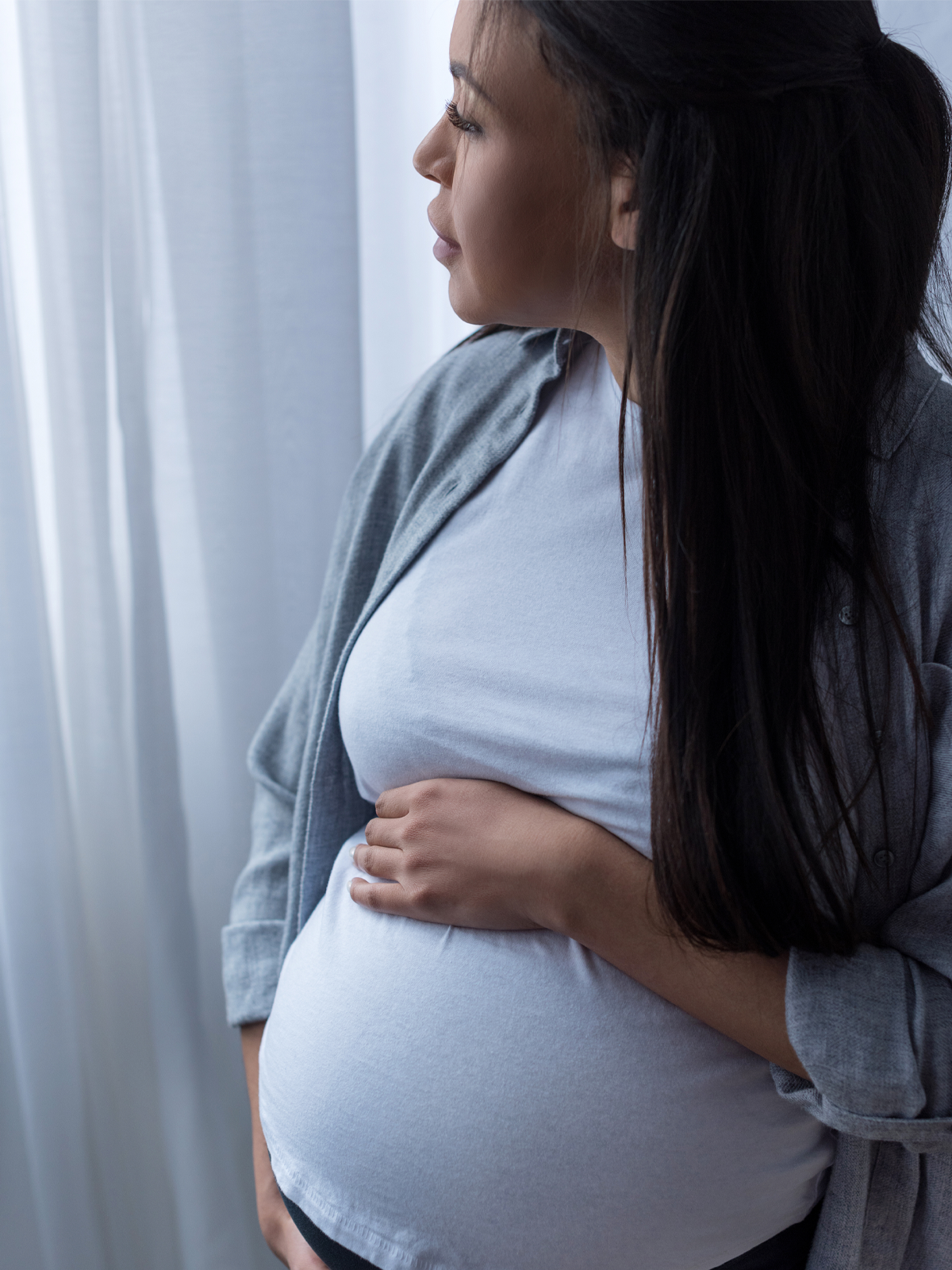 Pregnant woman gazing out the window, holding her belly.