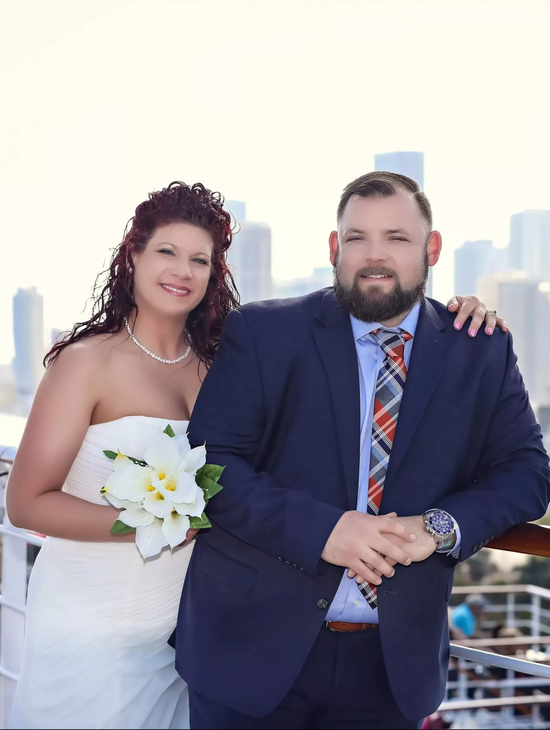 Newlyweds: Bride in strapless gown with bouquet, groom in navy suit, smiling, posed with cityscape background.