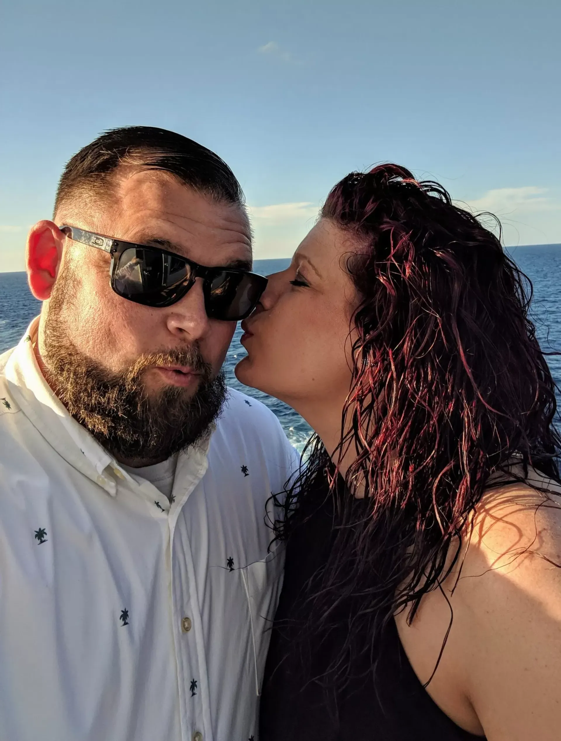 Woman kissing a bearded man wearing sunglasses on a boat, blue water and sky in background.