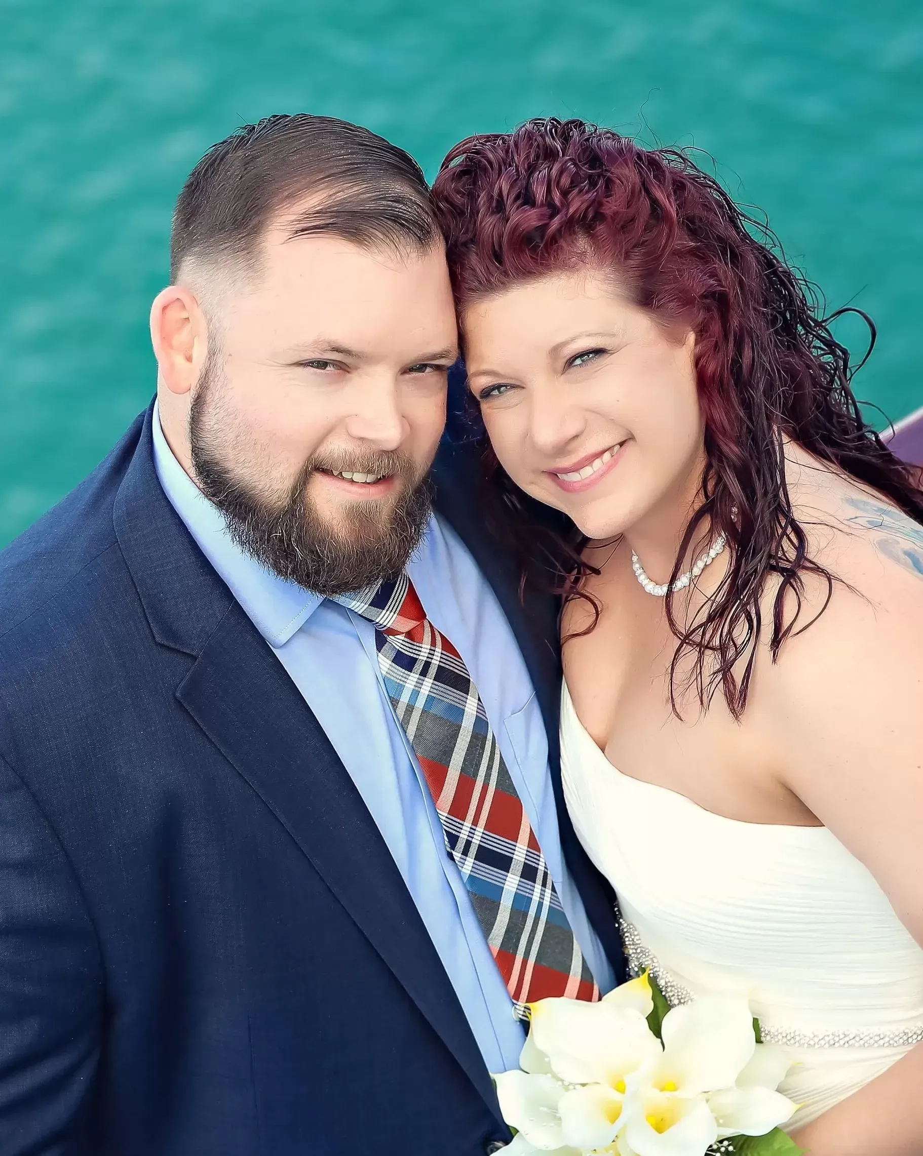 Couple in wedding attire; man with beard, blue suit, plaid tie; woman in white dress, red curly hair, smiling.