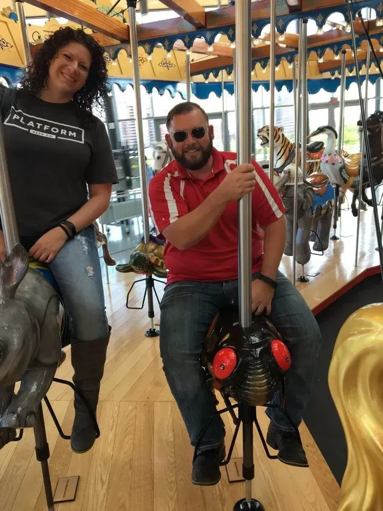 Woman and man smiling on a carousel, she in gray shirt, he in red. Indoor setting.