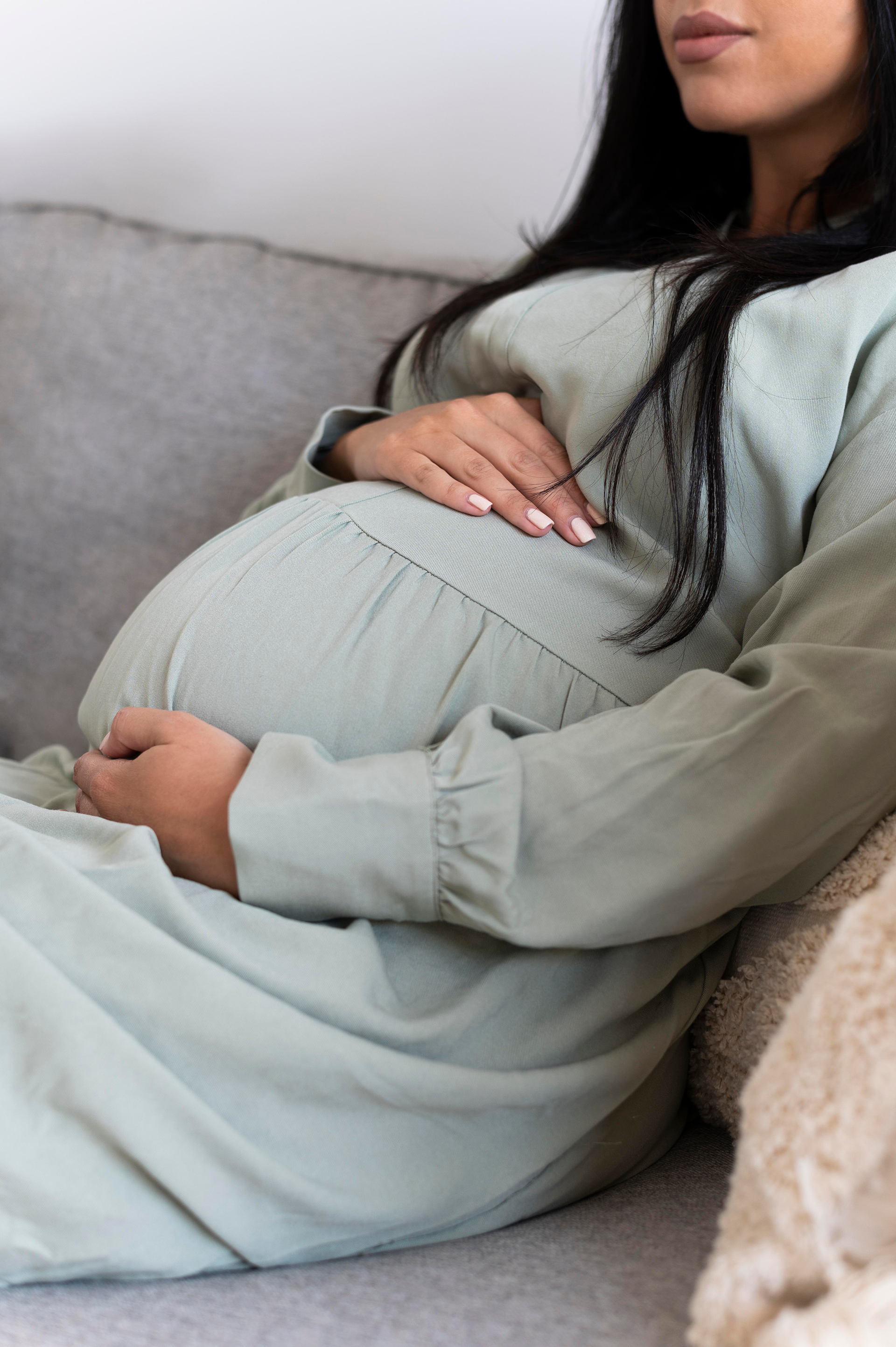 Pregnant woman in green dress sitting on a gray couch, hands on belly.