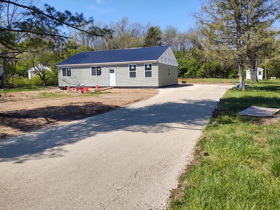 Gravel driveway leads to a light green house with a blue roof. Trees surround the property.
