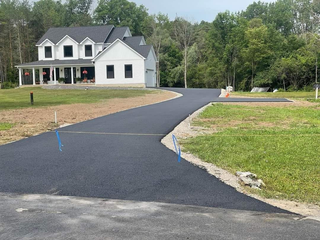 Newly paved asphalt driveway leading to a two-story white house with a porch; green grass and trees in the background.