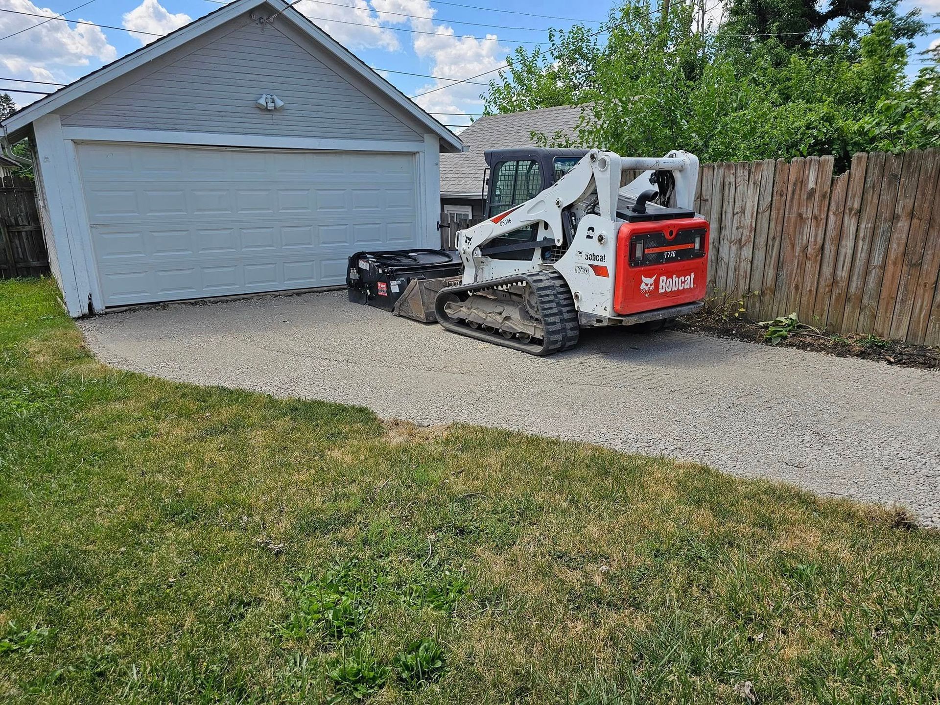 Bobcat skid steer grading gravel driveway near garage, alongside a wooden fence, on a sunny day.