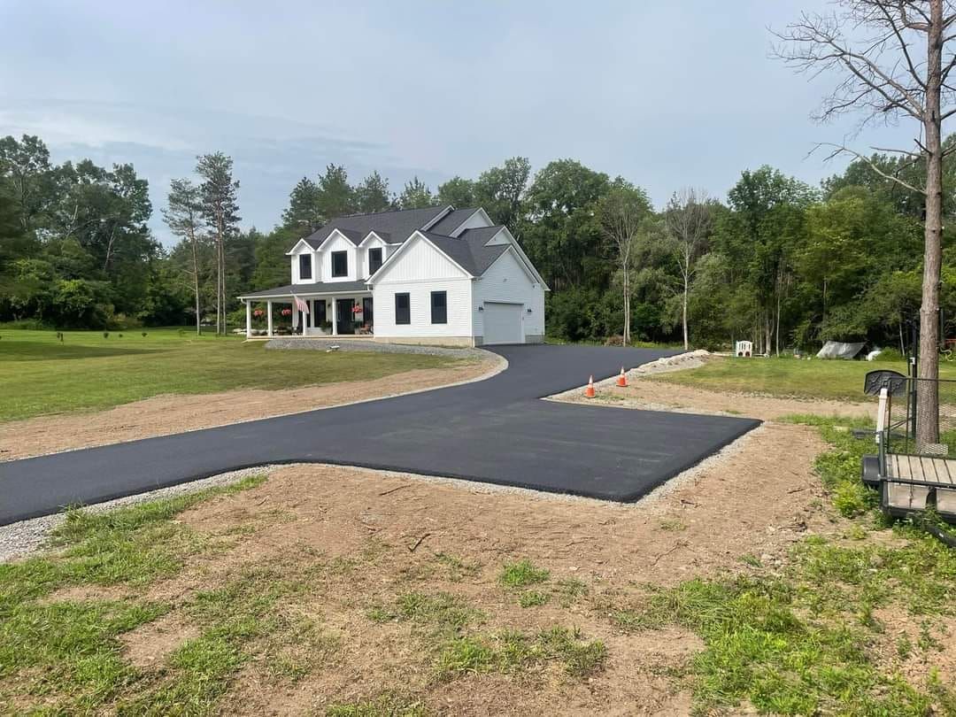 Black asphalt driveway leading to a white house with attached garage, set in a grassy, wooded area.