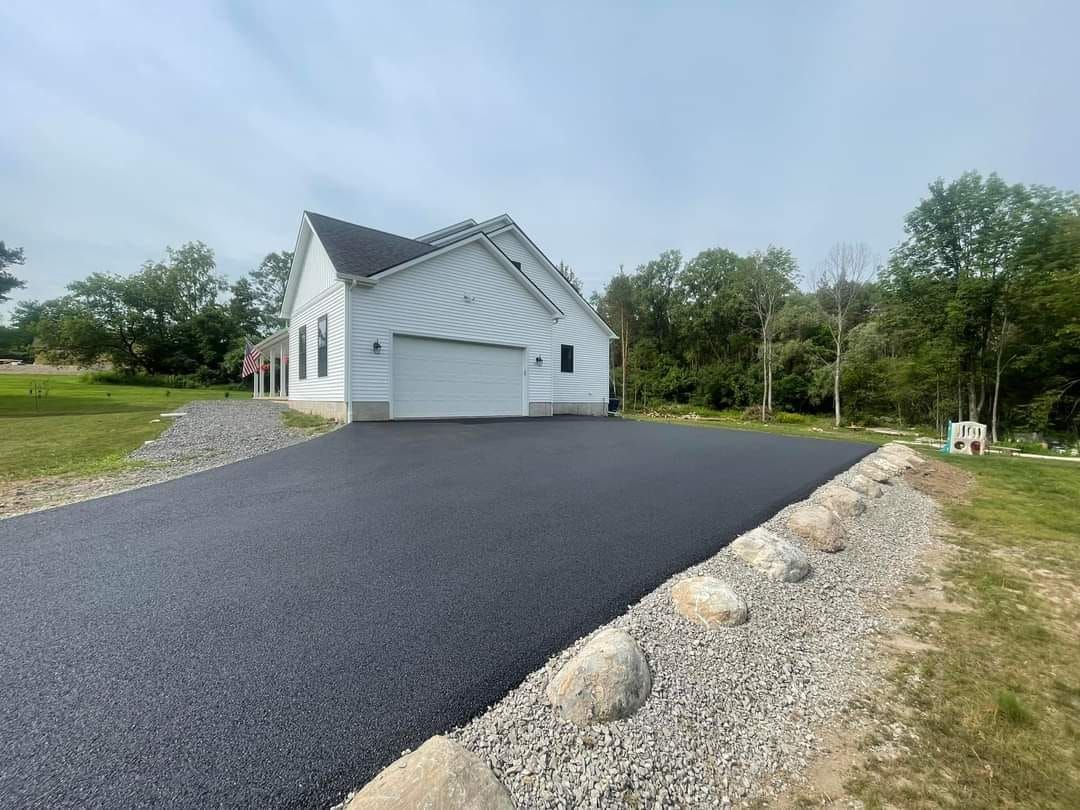 Asphalt driveway leading to white garage in a grassy area, lined with rocks.