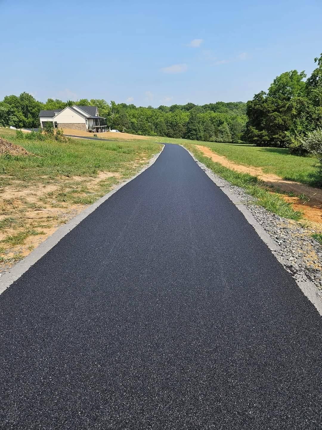 Asphalt path through a grassy area, leading towards trees and a house under a clear blue sky.