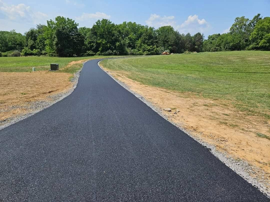 Black asphalt path curves through a grassy field, with trees in the background, on a sunny day.
