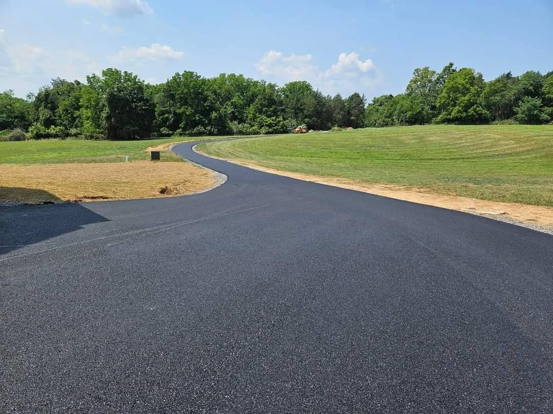 Dark asphalt driveway curves through a grassy field toward a tree line under a blue sky.