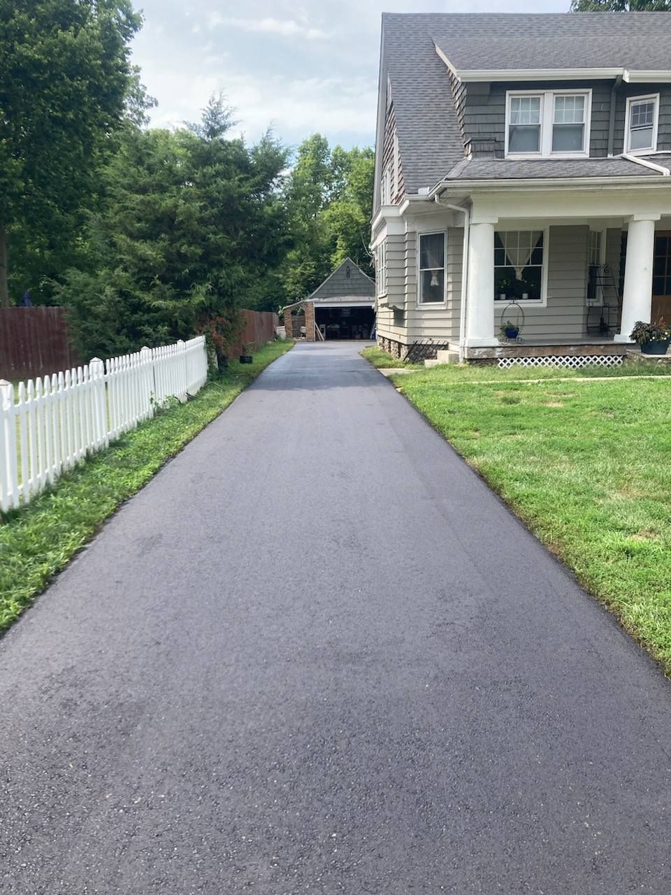 Newly paved asphalt driveway leading to a garage, with a white picket fence on the left and a house on the right.