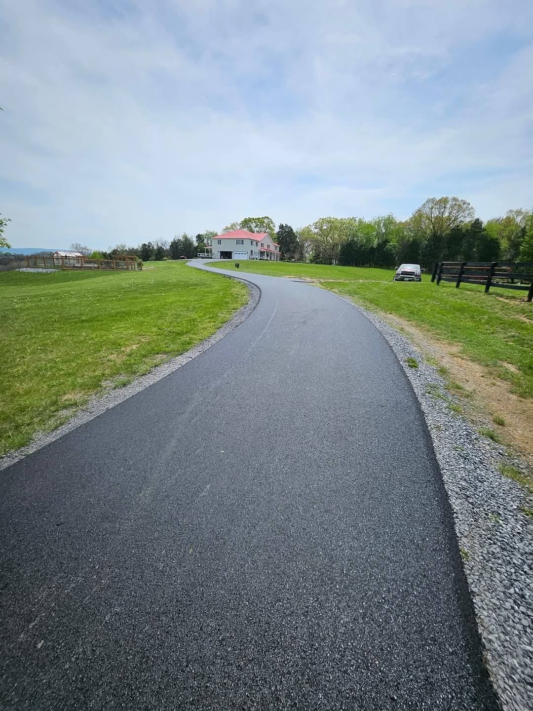 Paved path curves through a grassy field toward a house under a partly cloudy sky.