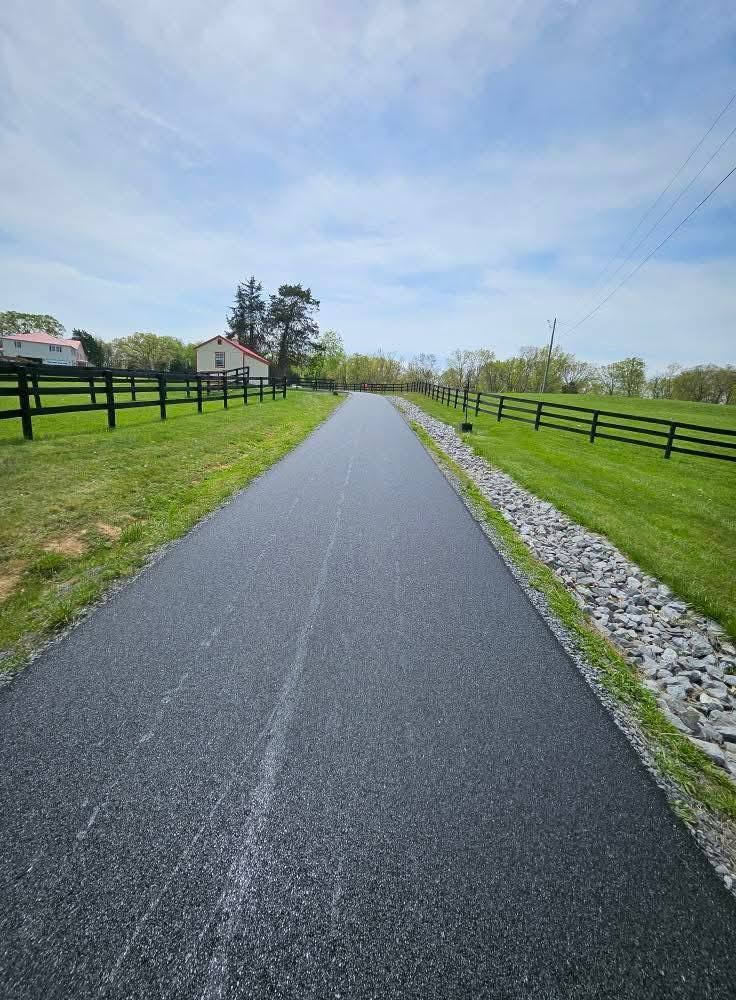Asphalt path bordered by green grass and black fences, leading to a distant house under a blue sky.