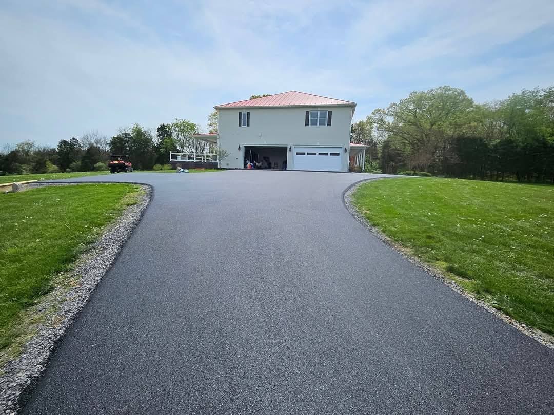 Asphalt driveway leading to a two-story house with a garage, flanked by green lawns under a blue sky.