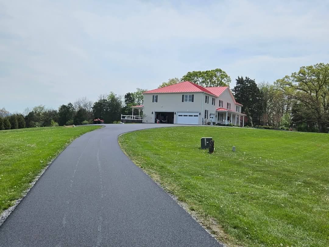 A long asphalt driveway leads to a two-story house with a red roof and a garage, set in a grassy yard.