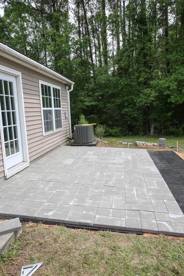 Gray paver patio next to a light-colored house with a door, window, and surrounded by trees.