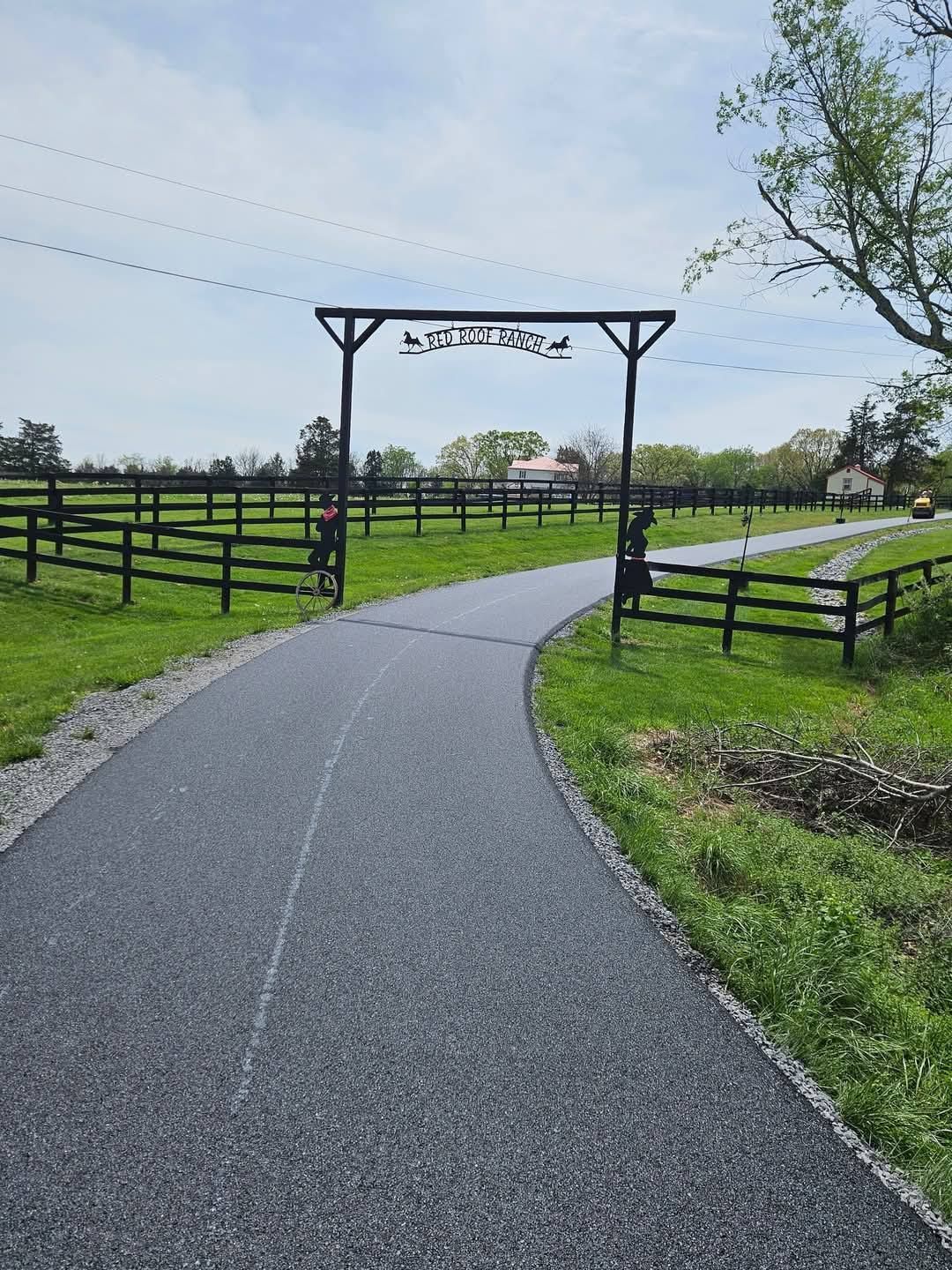 Black asphalt road leads to a farm entrance with sign and black fence.