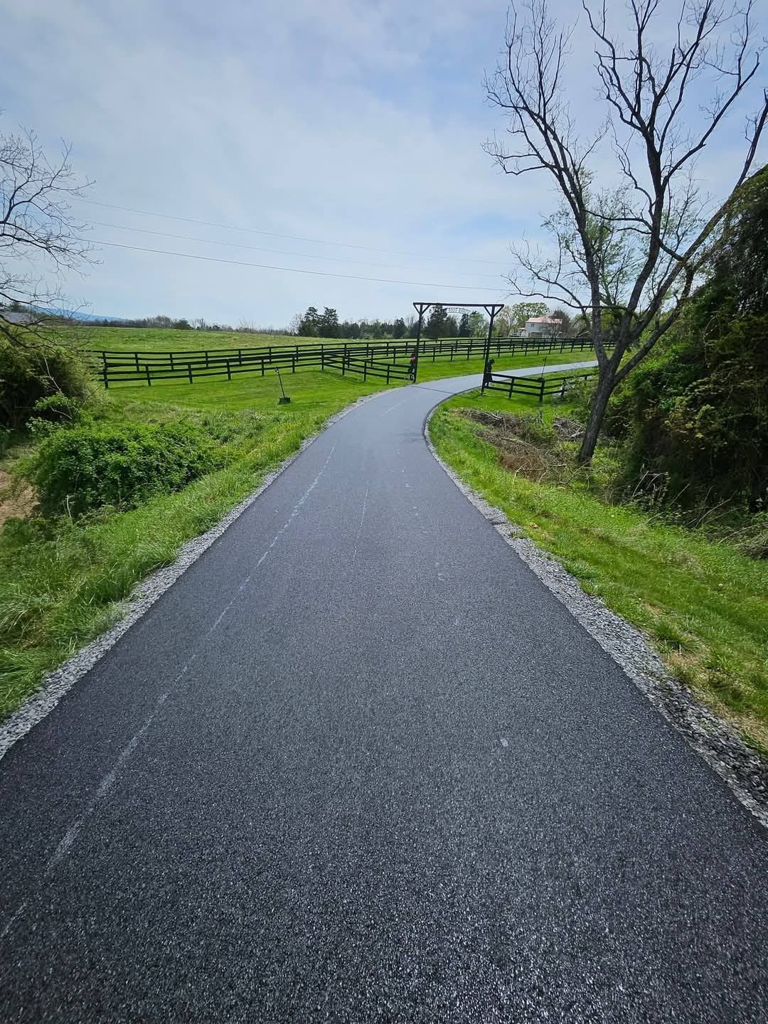 Paved path through green field, leading toward a fence, under a blue sky.