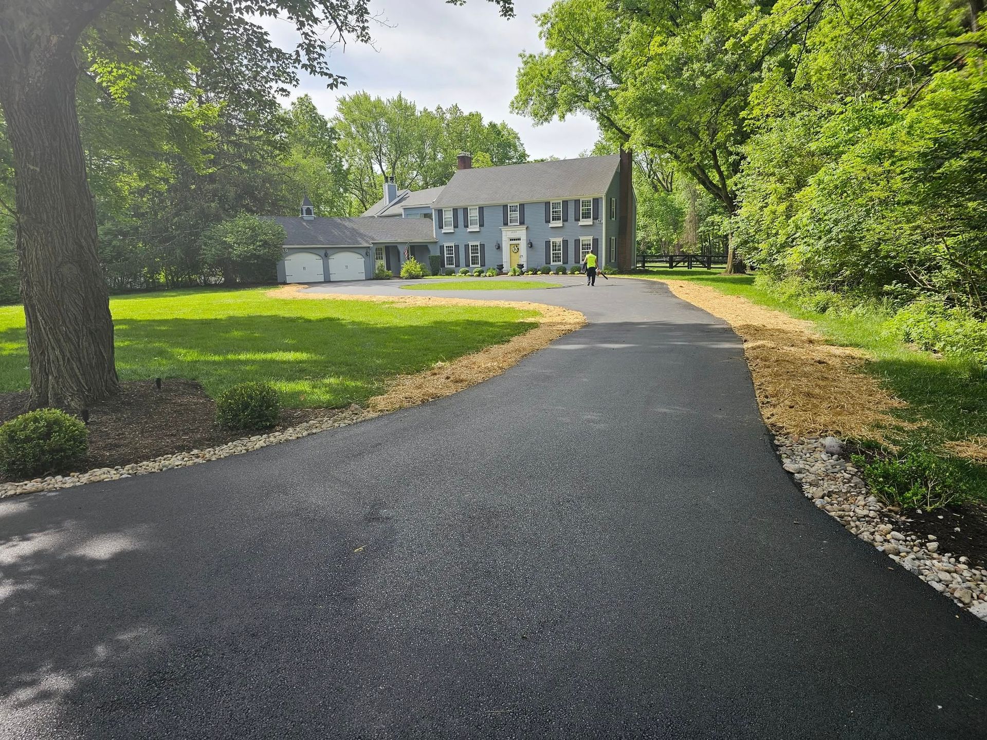 Black asphalt driveway leads to a blue house with green lawn and trees.