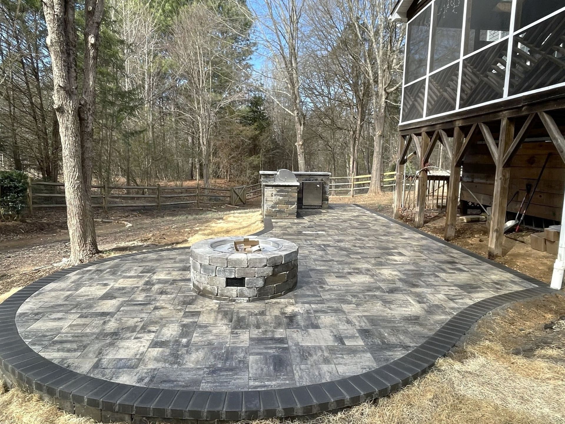 Circular brick patio with fire pit and outdoor kitchen; wooded background.