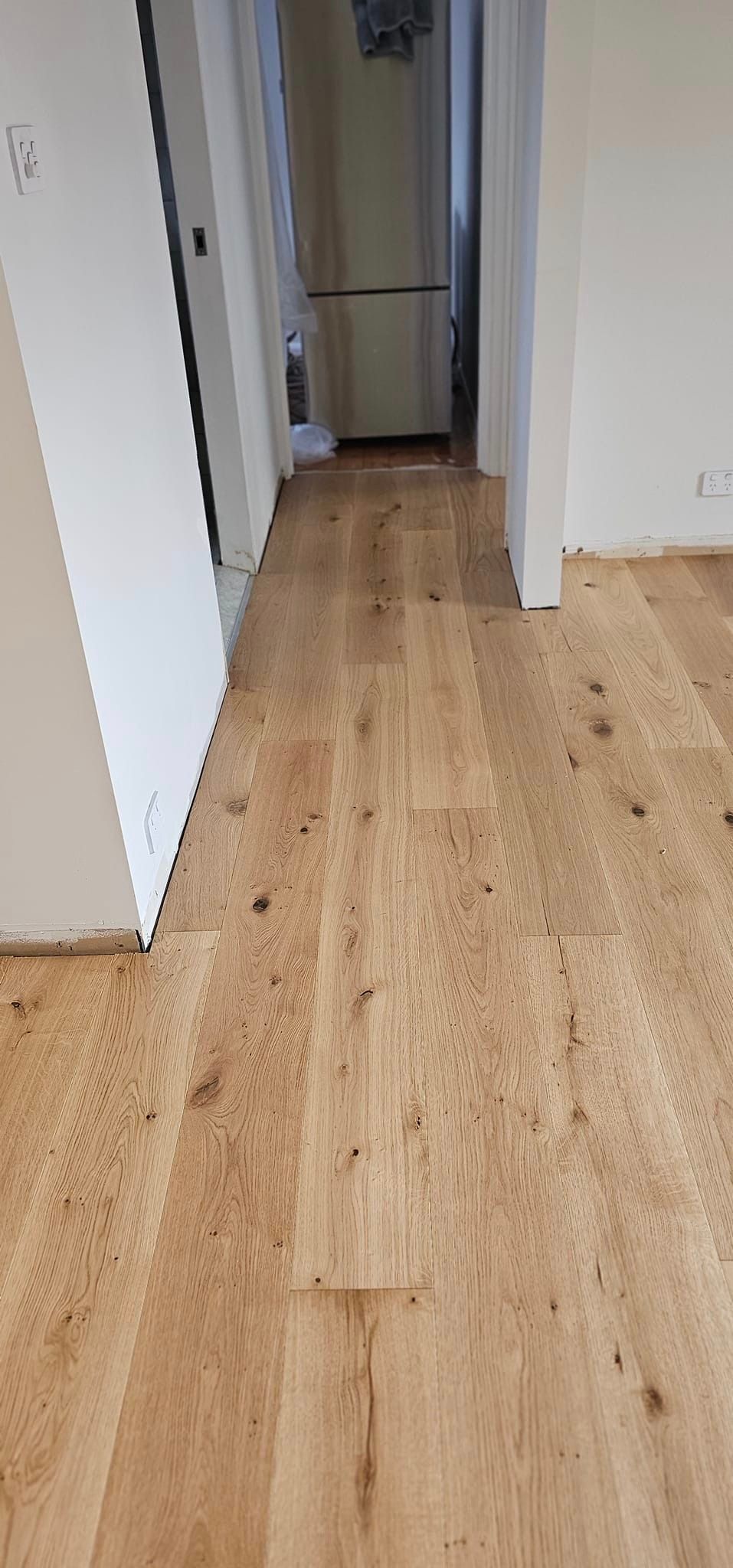 A hallway with a wooden floor and a refrigerator in the background.