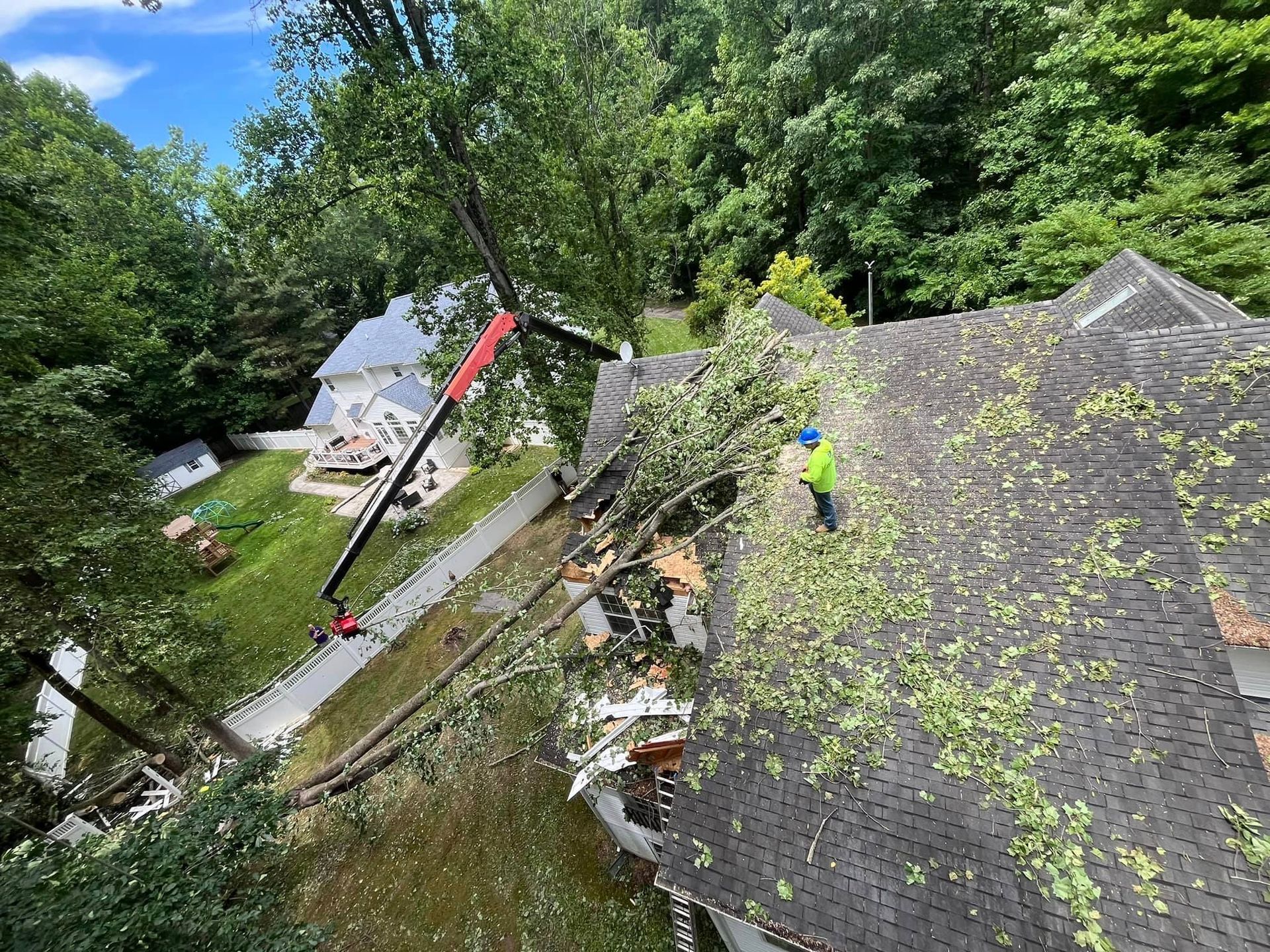 A man is standing on the roof of a house cutting a tree.